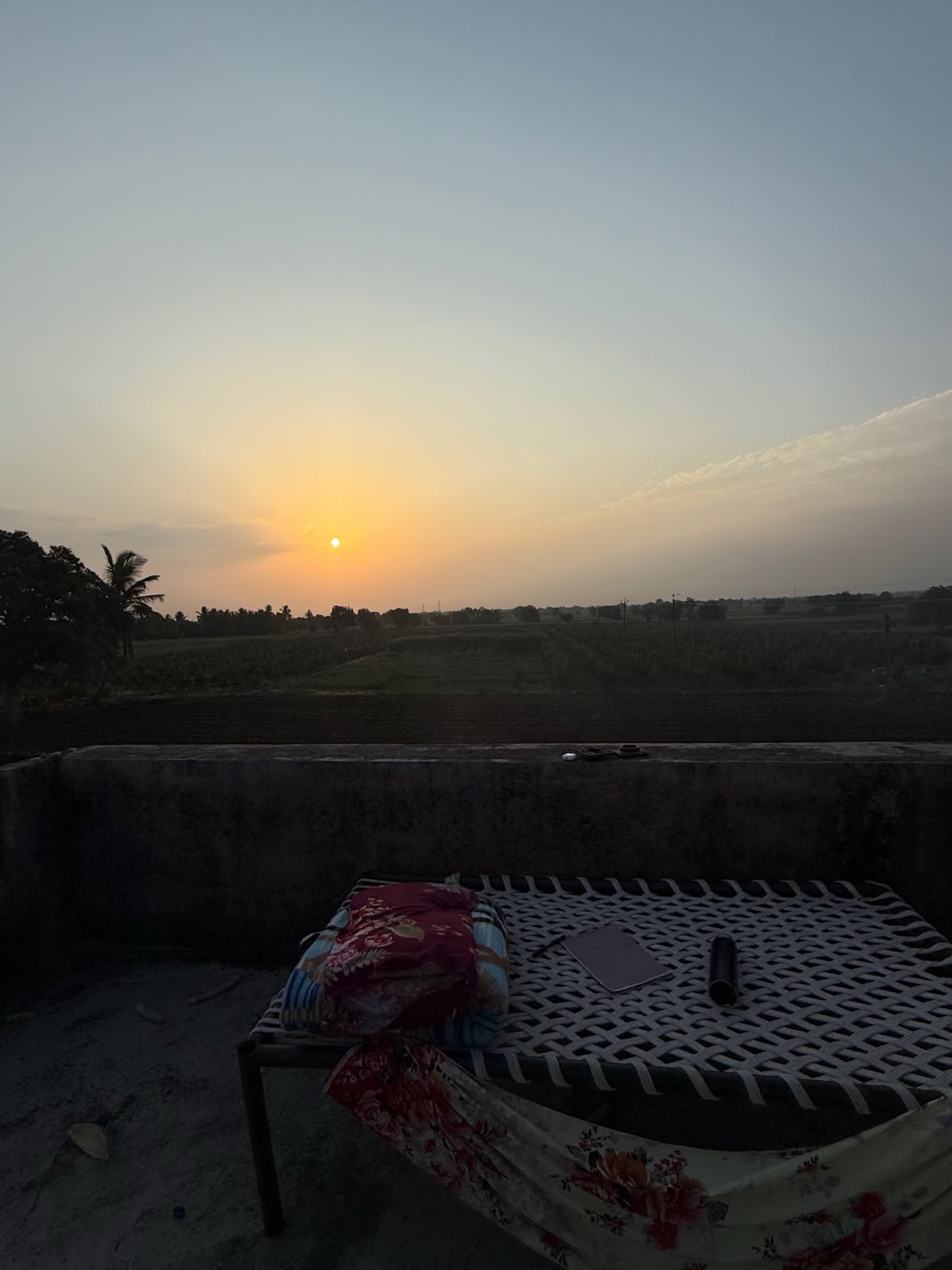 A rope cot with a pillow and an open notebook on a rooftop terrace, fields and a setting sun beyond.