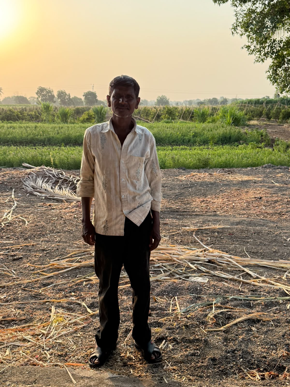 A man standing in a field at sunset, smiling gently, wearing a light shirt and dark trousers.